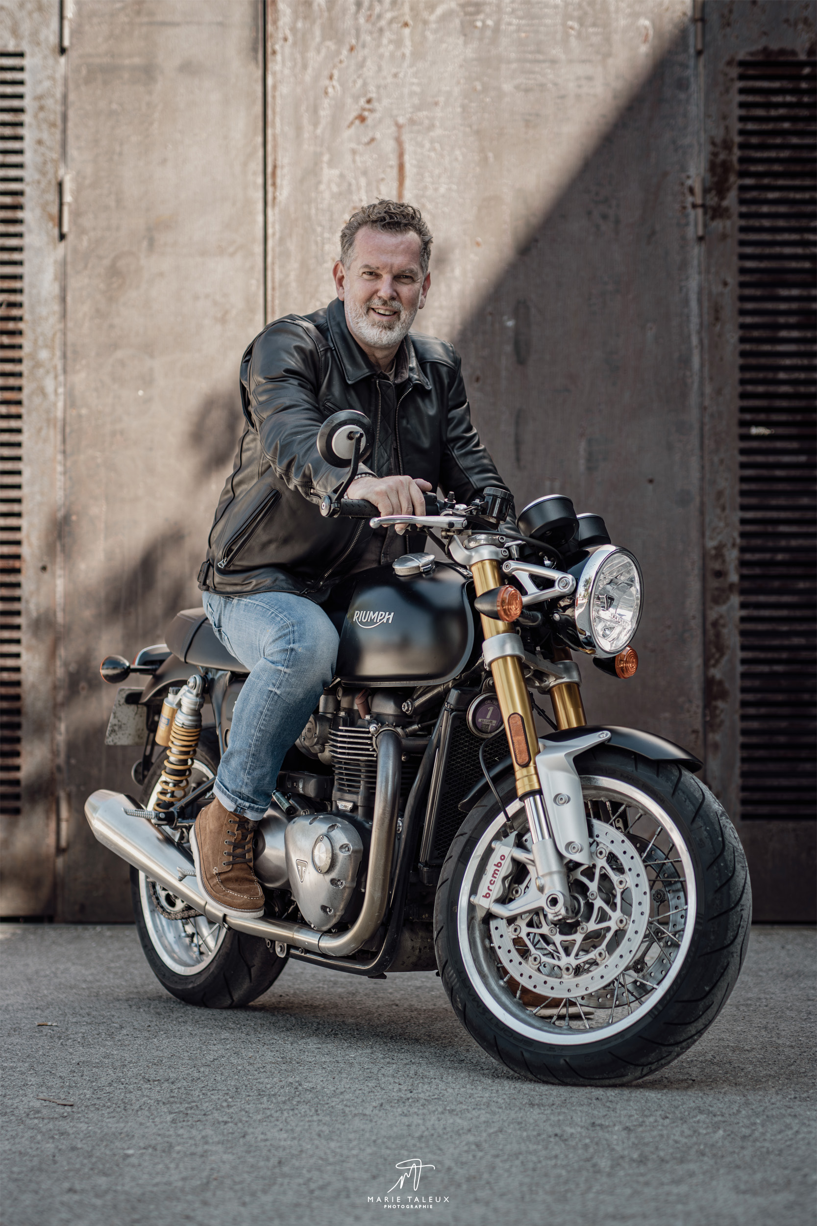 seance-photo-portrait-homme-moto-lyon-2 Portrait d’un homme assis sur une moto Triumph noire devant un décor industriel à Lyon.