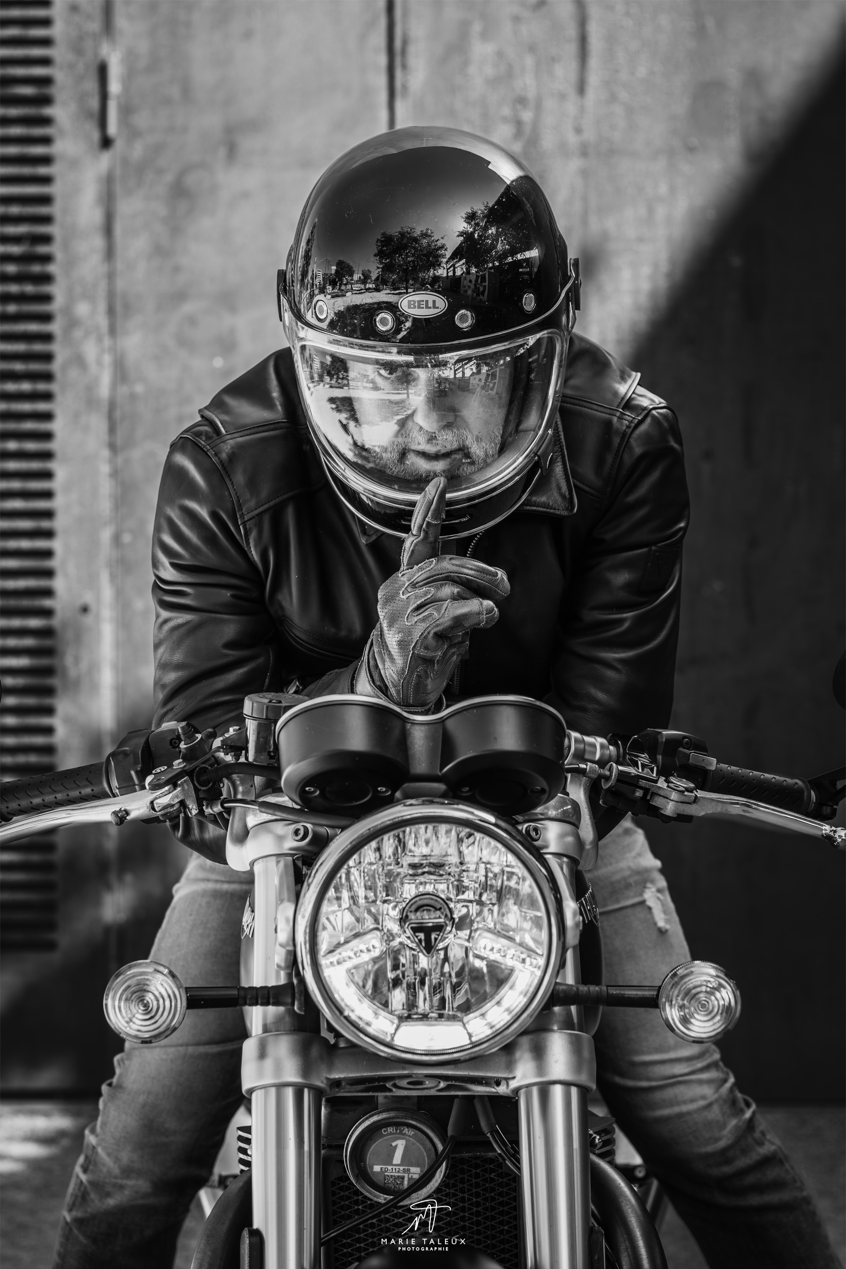 seance-photo-portrait-homme-moto-lyon-3 Portrait en noir et blanc d’un motard casqué face à l’objectif, installé derrière sa moto Triumph.