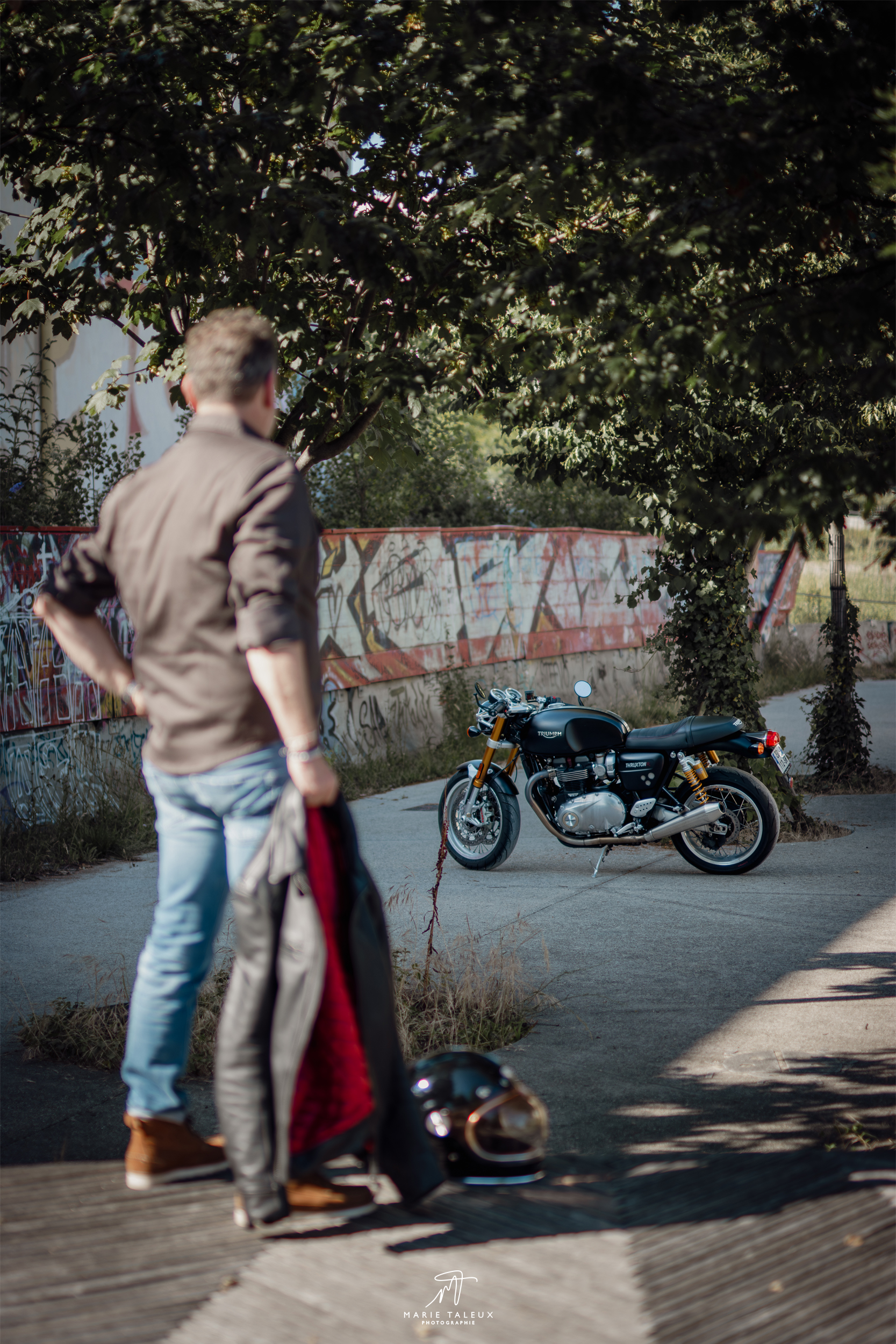 seance-photo-portrait-homme-moto-lyon-4 Motard vu de dos marchant vers sa moto Triumph noire dans un environnement urbain et végétalisé.