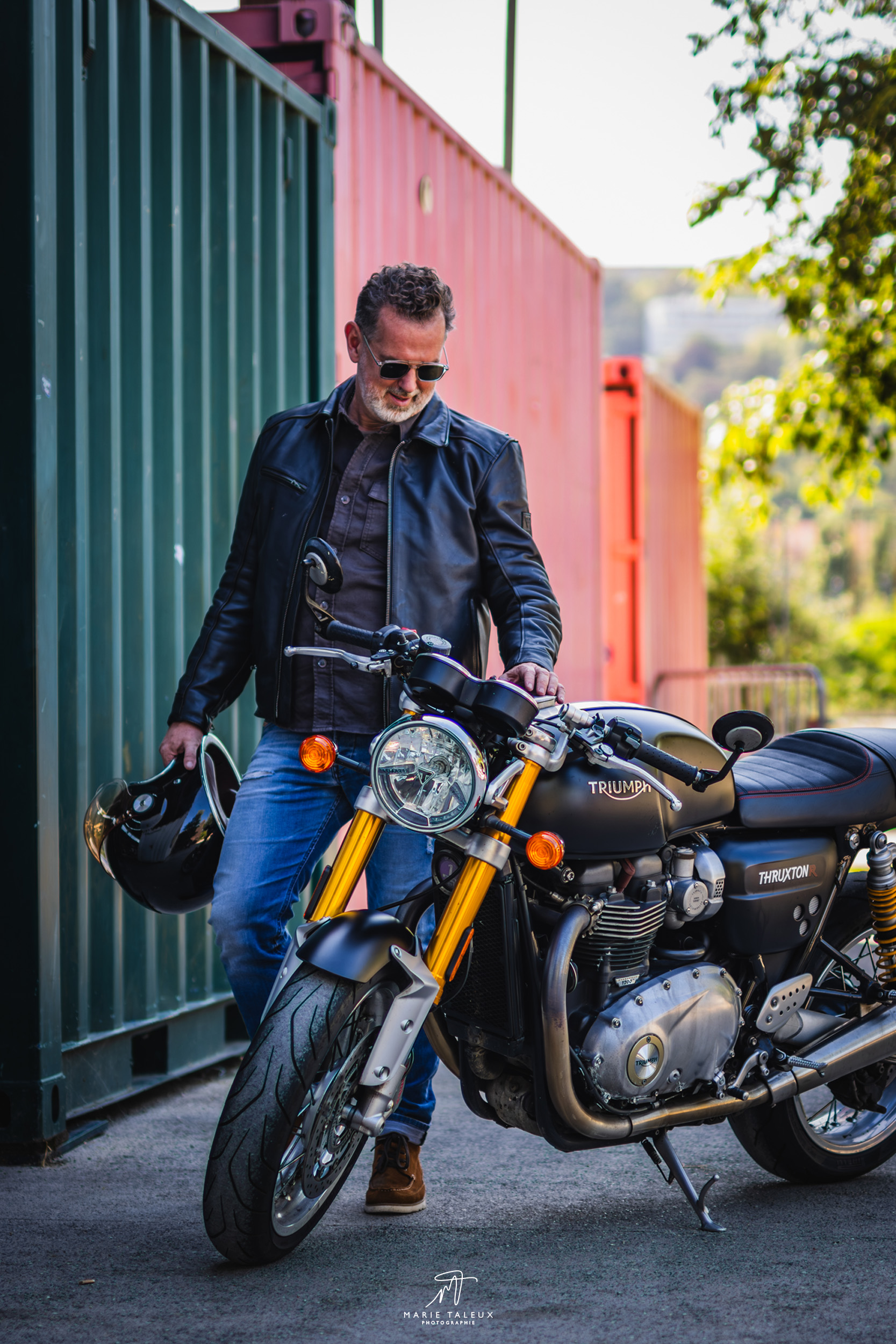 seance-photo-portrait-homme-moto-lyon-5 Portrait d’un homme en veste en cuir debout à côté de sa moto Triumph noire, dans un décor de containers colorés.