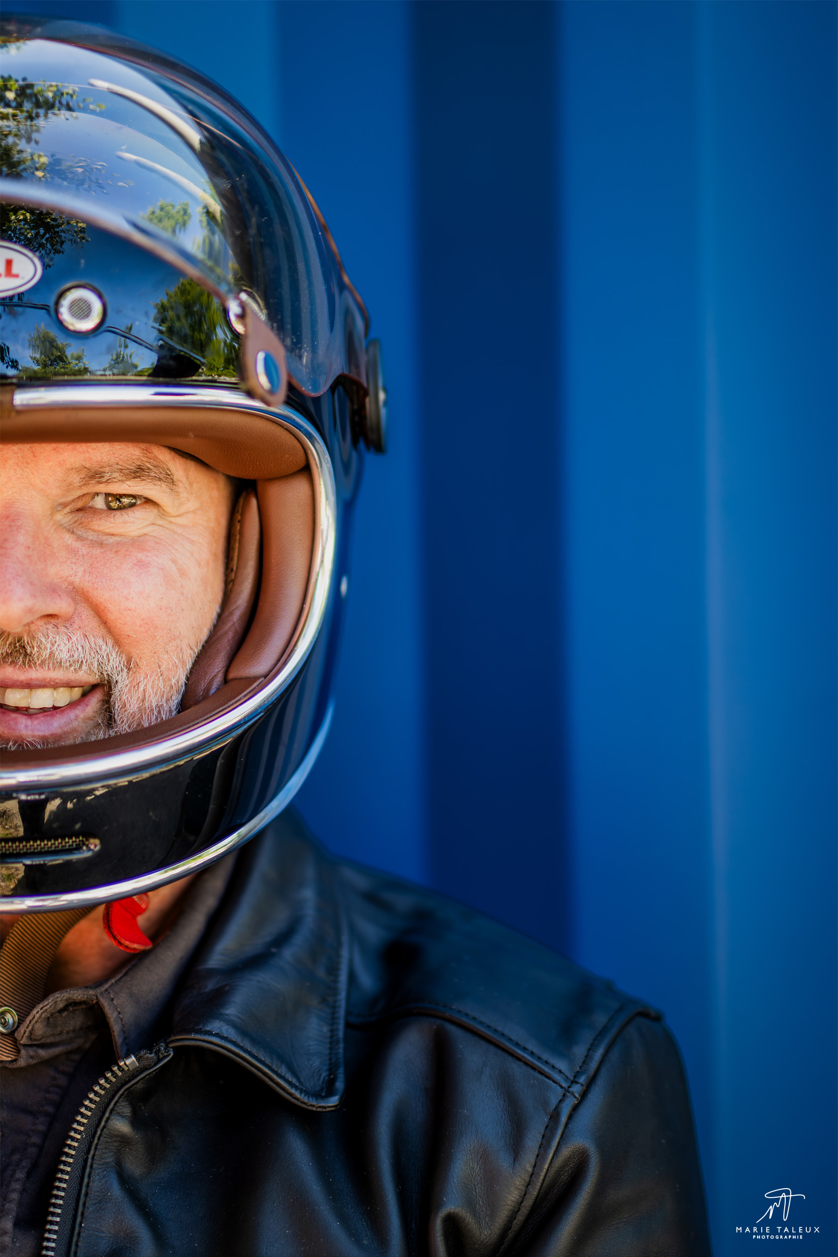 seance-photo-portrait-homme-moto-lyon-6 Portrait d’un motard portant un casque noir et une veste en cuir, photographié de faceà Lyon, sur fond bleu.