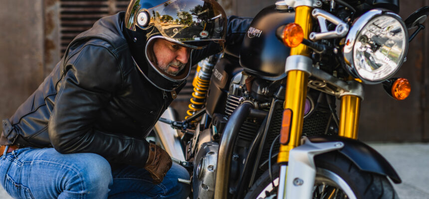 Motard accroupi à côté de sa moto Triumph noire, dans une ambiance urbaine et industrielle.