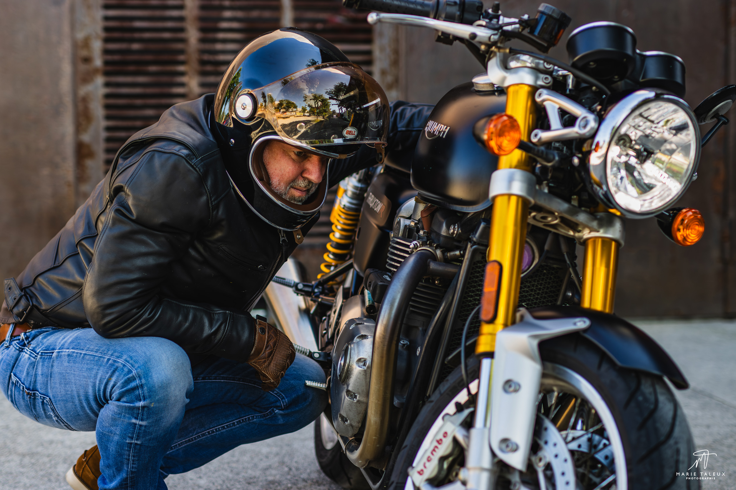 seance-photo-portrait-homme-moto-lyon-7 Motard accroupi à côté de sa moto Triumph noire, dans une ambiance urbaine et industrielle.