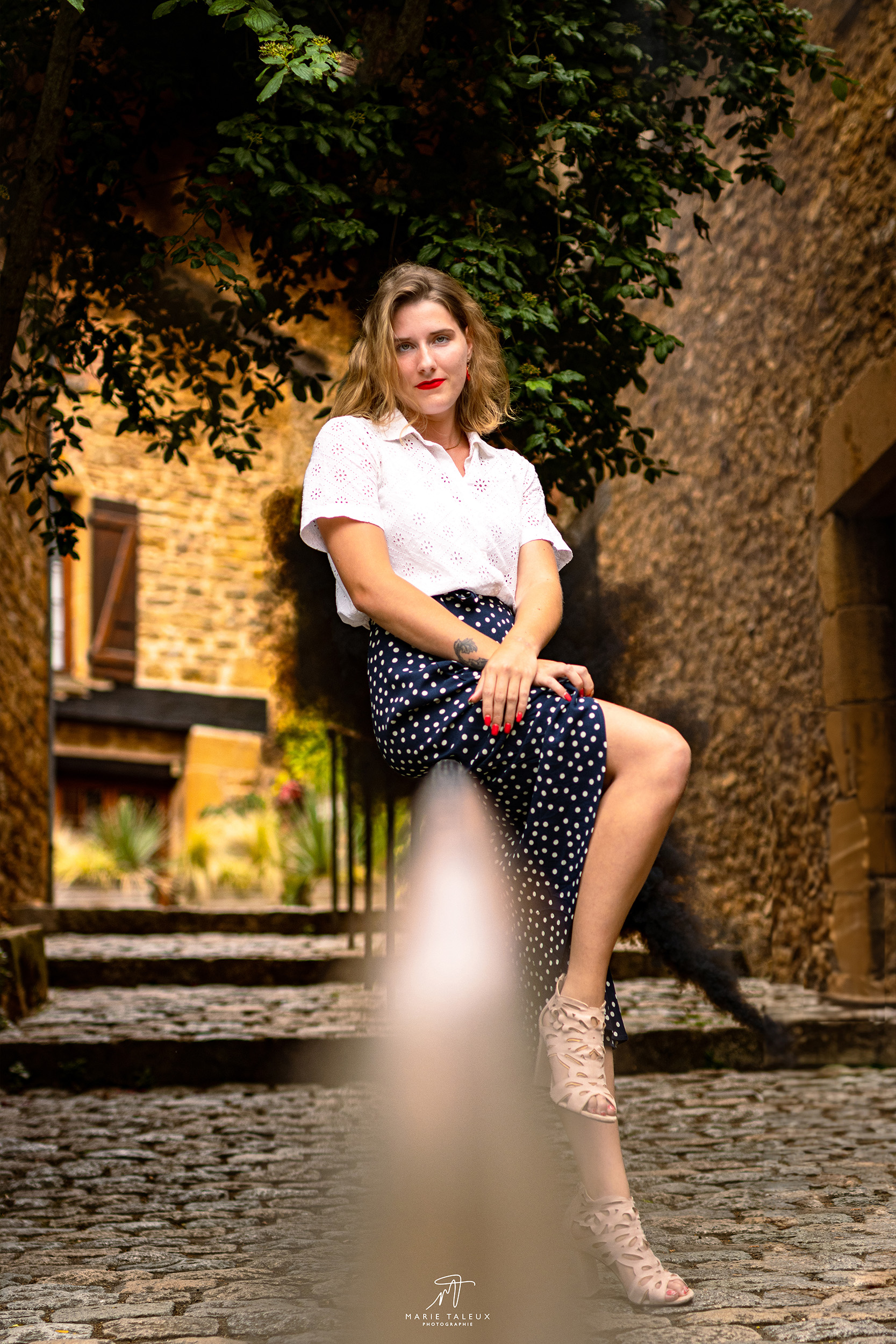 shooting-photo-portrait-femme-rhone-2 Jeune femme blonde assise dans une ruelle pavée, vêtue d’un chemisier blanc et d’une jupe longue bleu marine à pois, posant devant un mur en pierre et de la végétation.