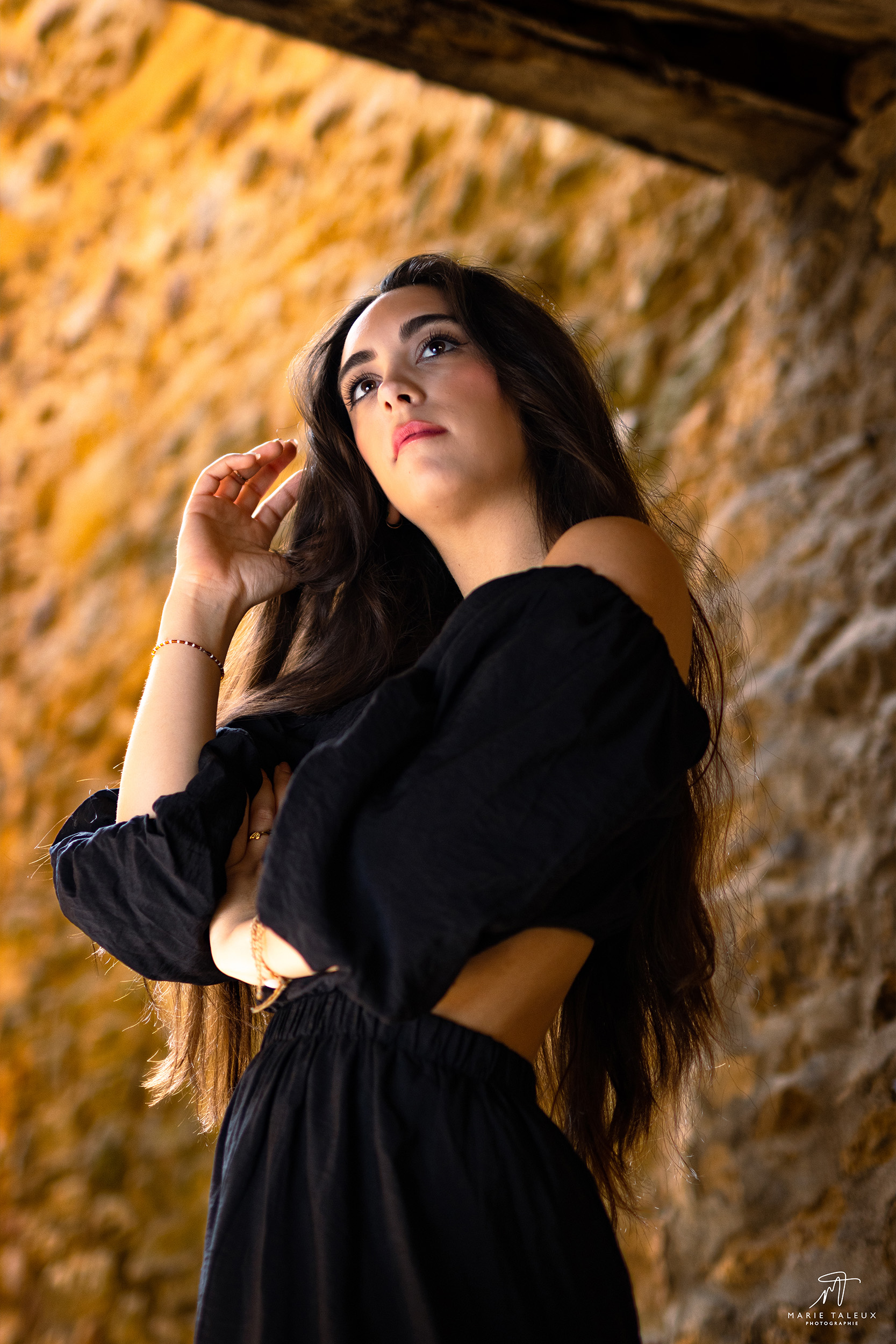 shooting-photo-portrait-femme-rhone-5 Portrait en contre-plongée d’une jeune femme brune en robe noire, regardant vers le haut sous une arche en pierre éclairée par une lumière chaude.